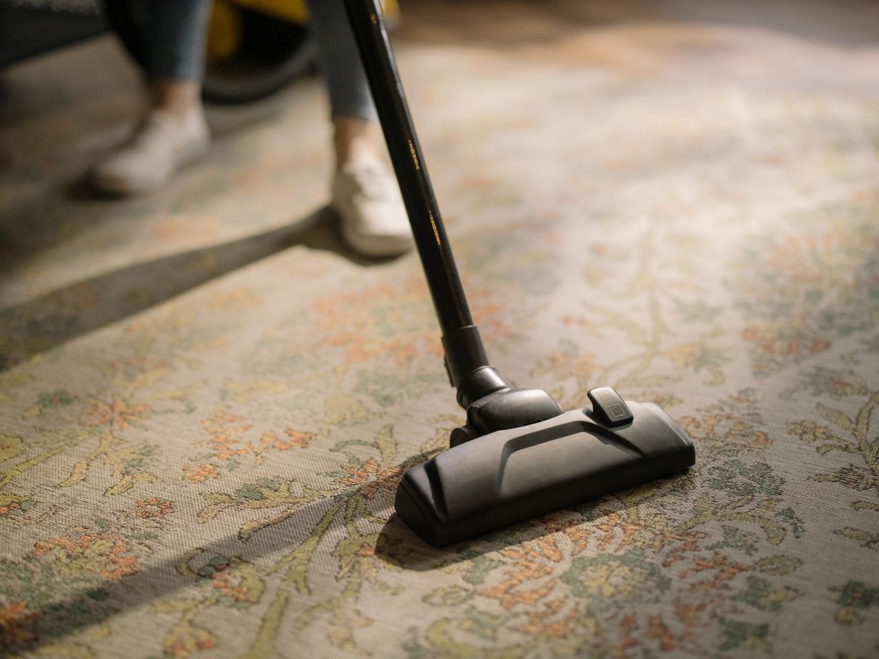 hero-img-01 Close-up of a vacuum cleaner on a patterned carpet in a sunlit room, capturing a moment of household cleaning.
